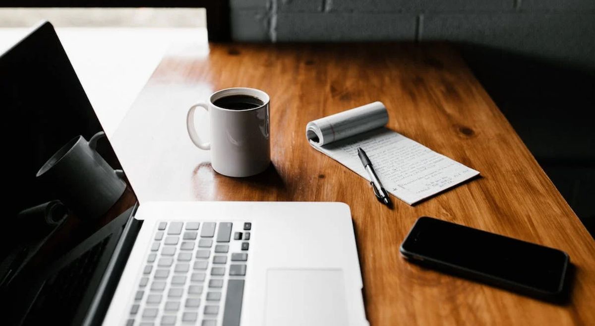 A picture of a computer on a desk alongside a mug, pen, paper and phone